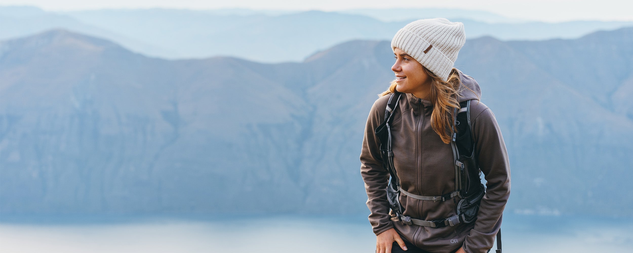 Female hiking in the mountains above the water wearing Outdoor Research Women's Crescent Fleece Full Zip Hoodie Sable and Liftie VX Beanie Sand.