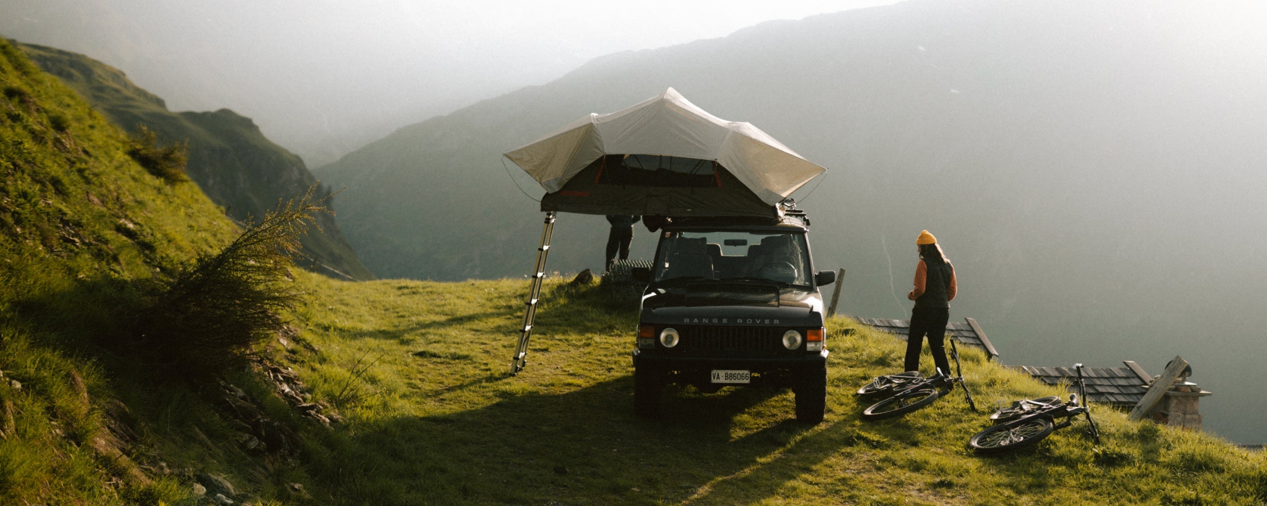 A woman walks around her truck camper on a green hillside.