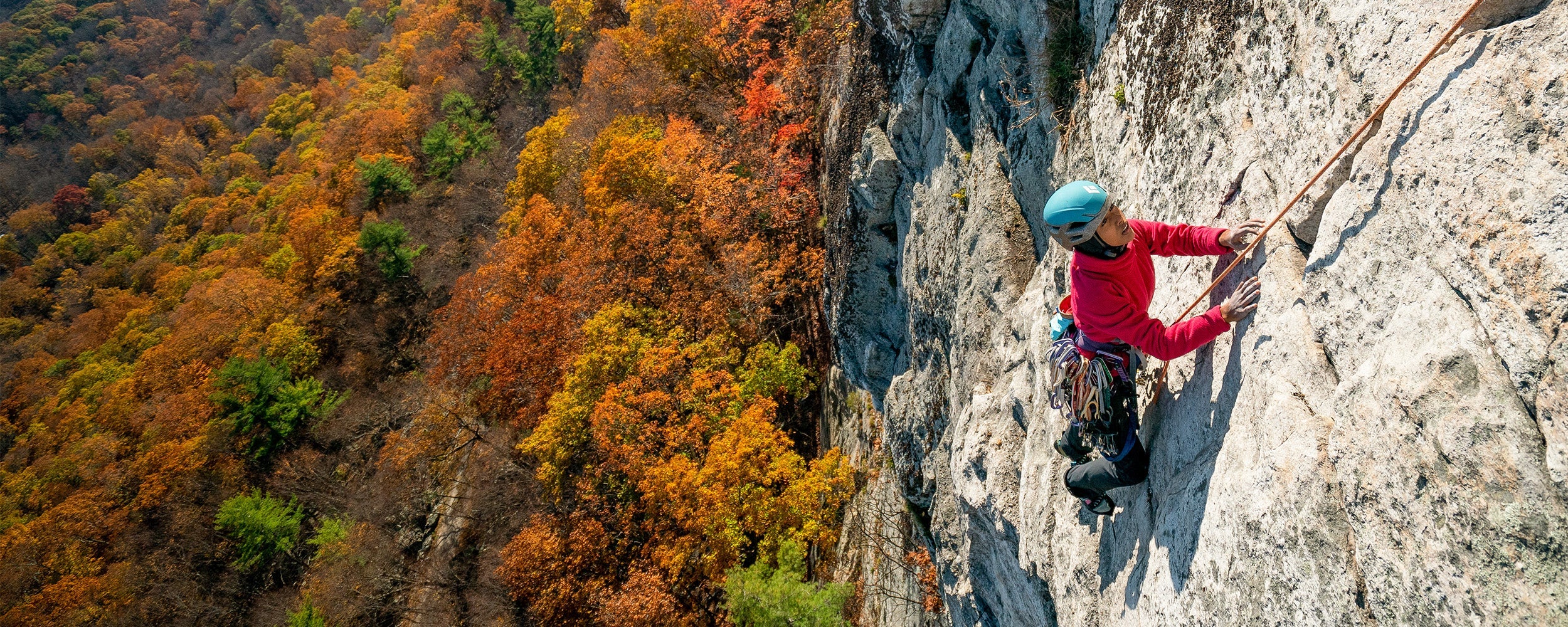 Person rock climbing on a rocky cliff face with autumn foliage in the background.