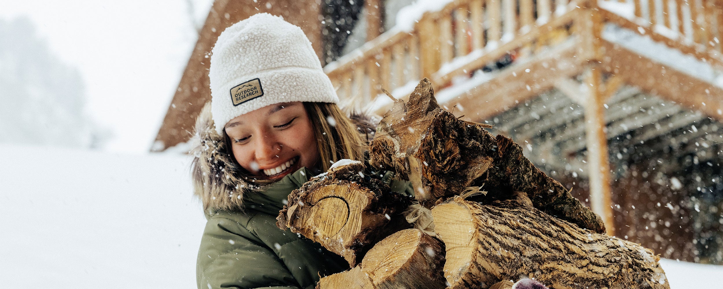 Zoe Atkin wears the Outdoor Research Grayland Fleece Beanie in Oyster while carrying fire wood in the snow. 