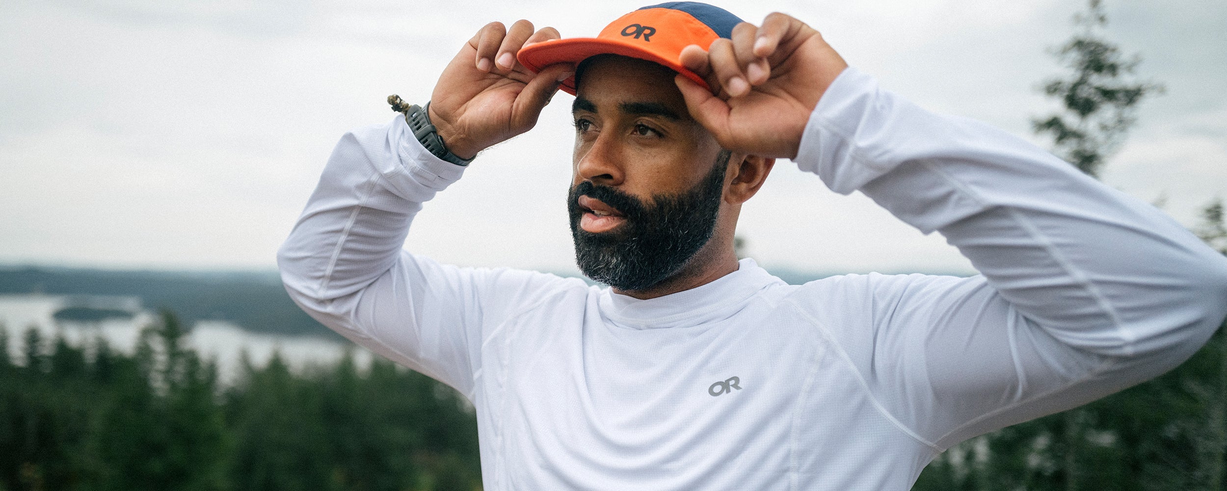 Man holds the brim of his Outdoor Research Swift Air Cap in Spice/Cenote as he gazes at the ocean views at the top of a hike. 