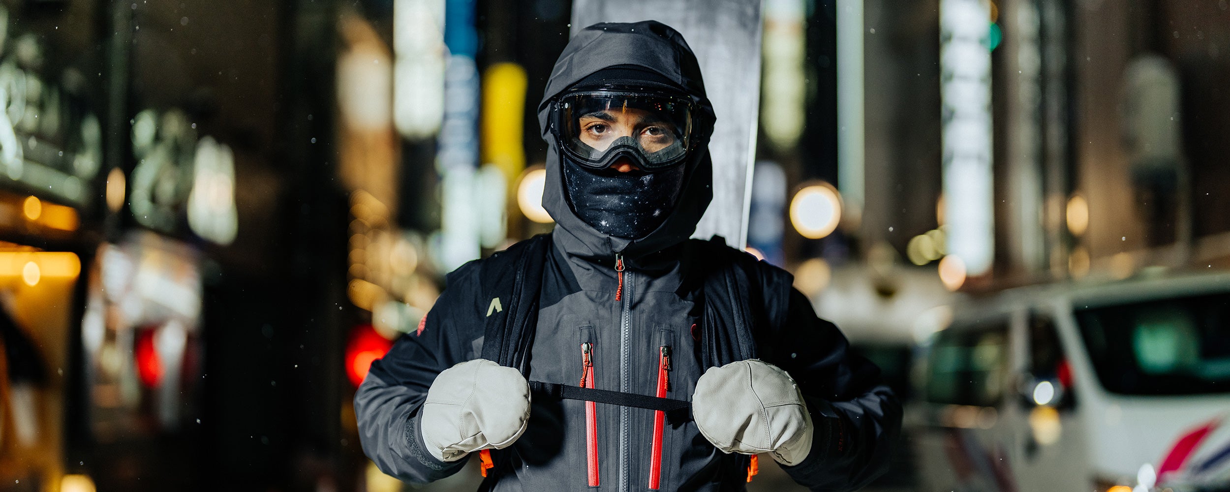 Man standing in a snowy city at night holds the straps of his ski backpack wearing a black balaclava. 