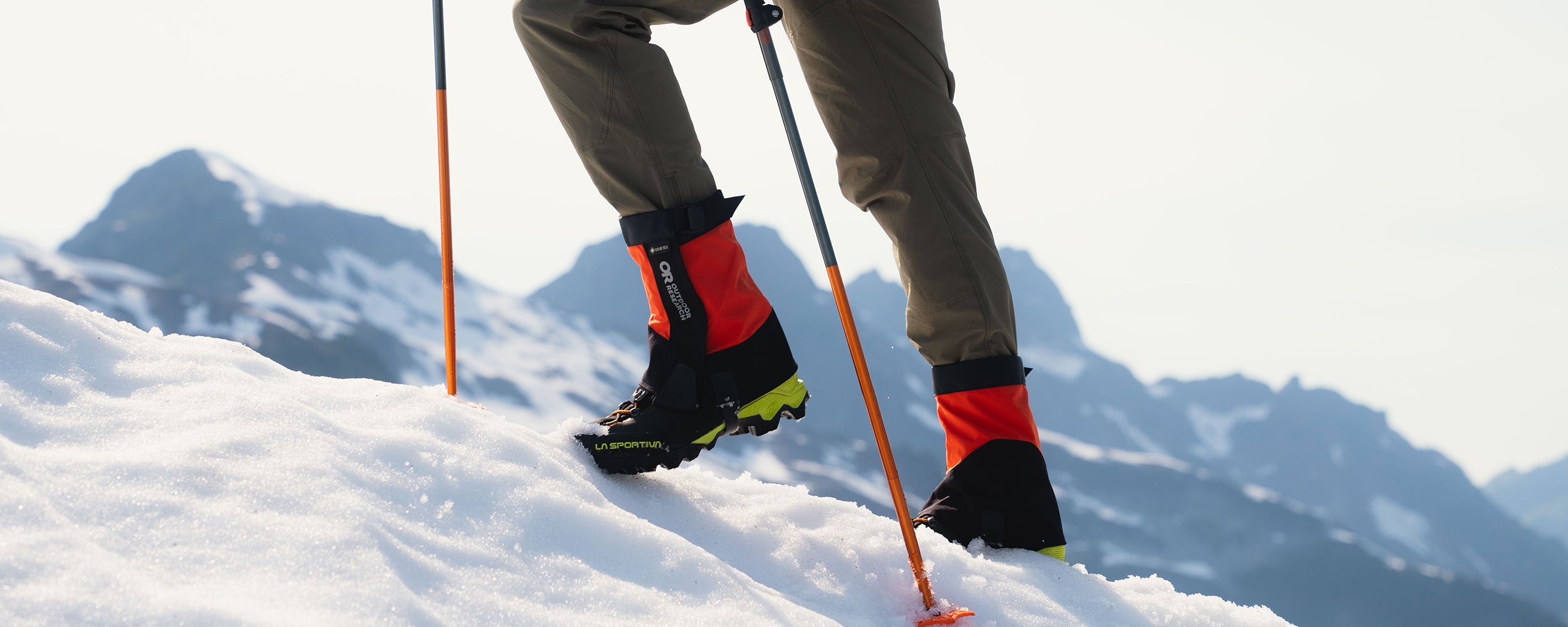  Man climbs up a snowy hill while wearing the Crocodile Mid-Height Gaiters in Spice/Black. 