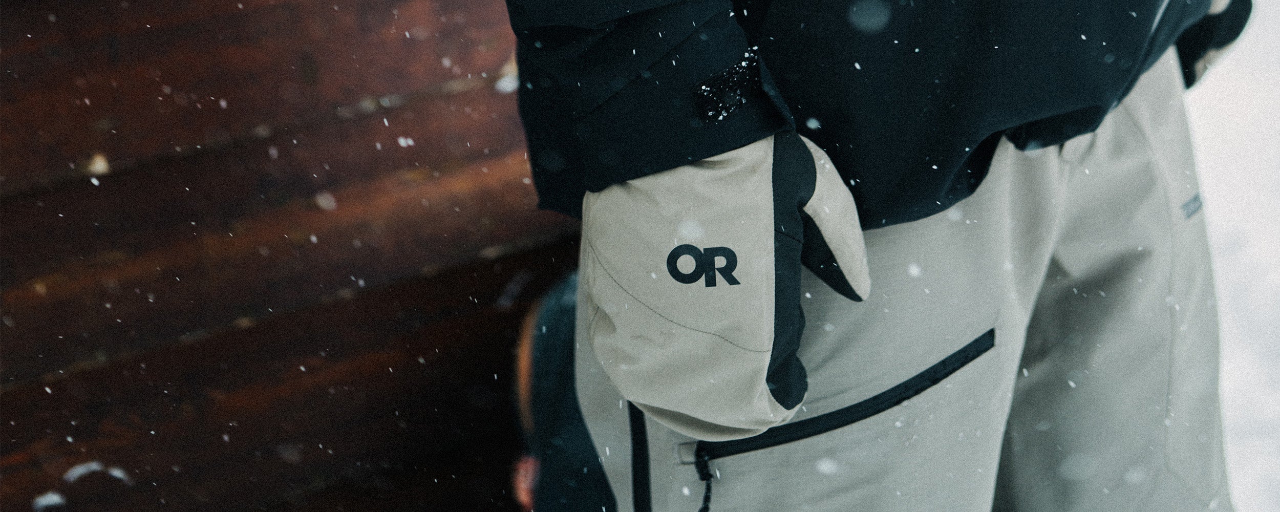 Male standing by the wall looking out at the snow wearing Outdoor Research Men's Powderverse Jacket Black and Hemispheres Team GORE-TEX 3L Mitts Sable. 