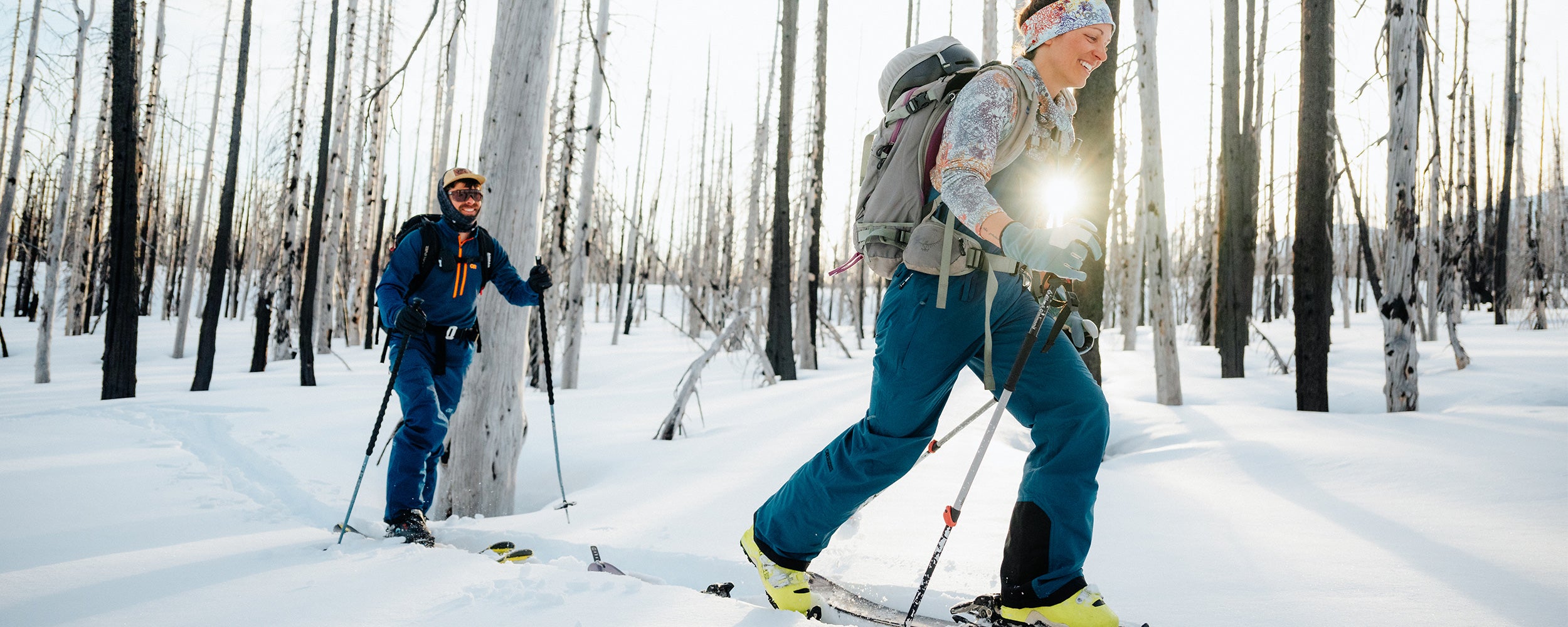 Two friends on a ski tour traverse through the trees wearing Outdoor Research Sportswear and Outerwear. 