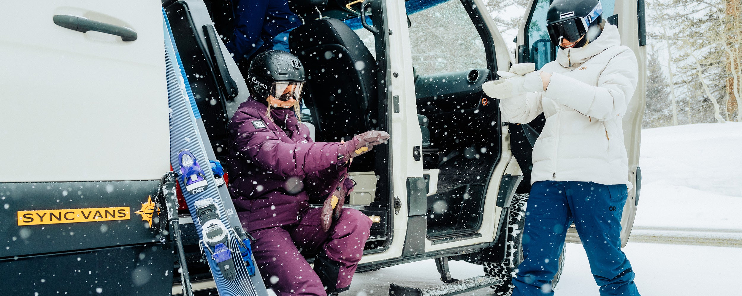 Two Females sit outside their Van in the snow to gear up in their Outdoor Research ski Outerwear. 