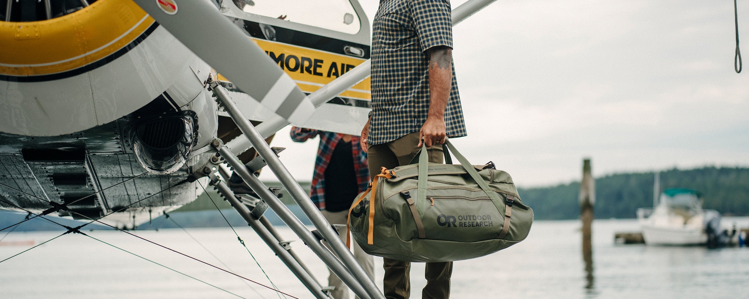 Man begins to load his Outdoor Research duffel bag onto a float plane while wearing the Astroman Short Sleeve Sun Shirt in Gold Nebula Plaid. 