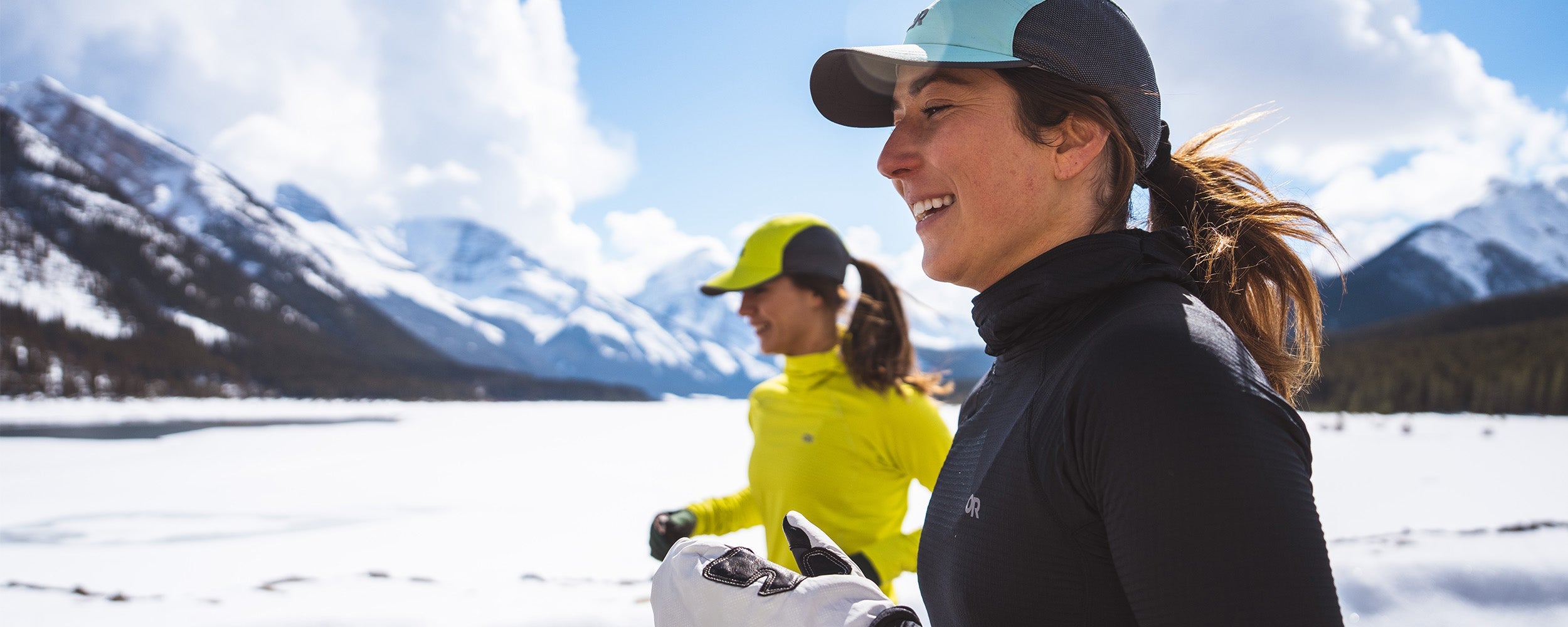 Two women run through snow wearing the Outdoor Research Vigor Grid Fleece Pullover Hoodie in Black.