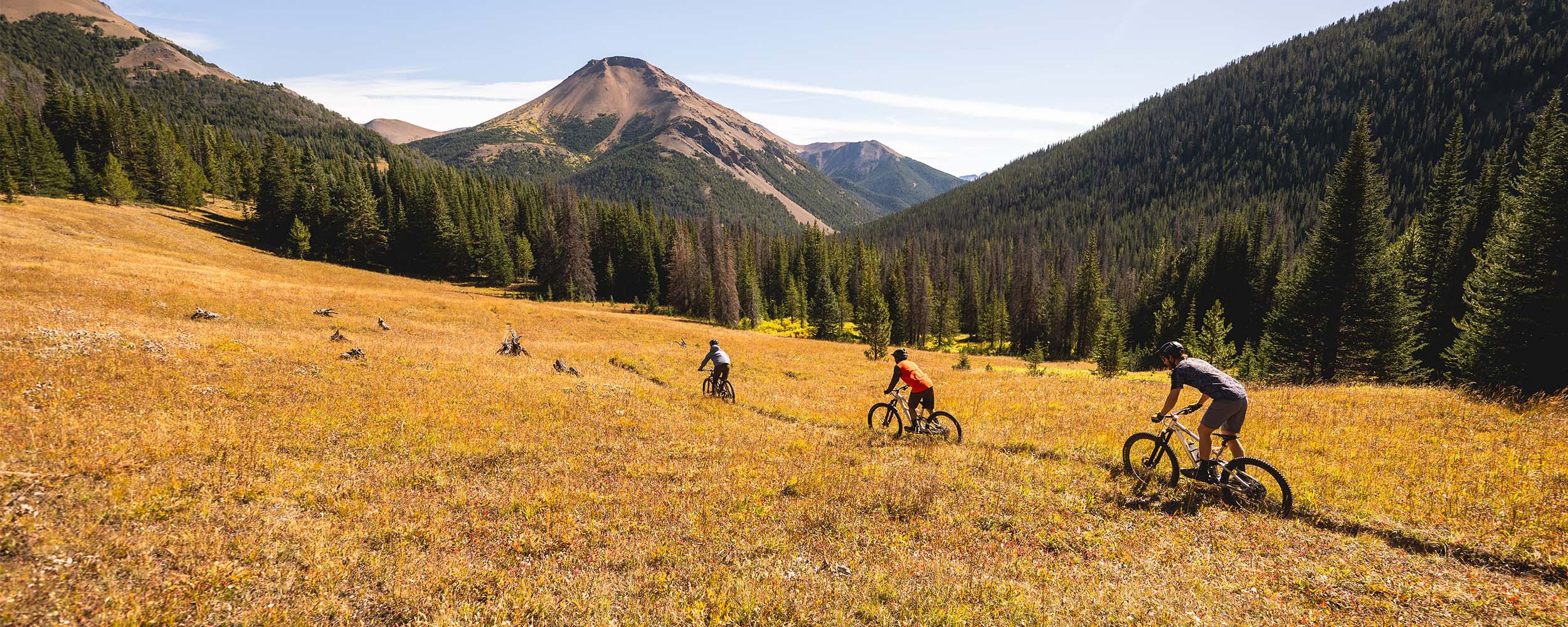 Three mountain bikers wearing Outdoor Research Sportswear ride along a dirt path beneath the blue sky. 