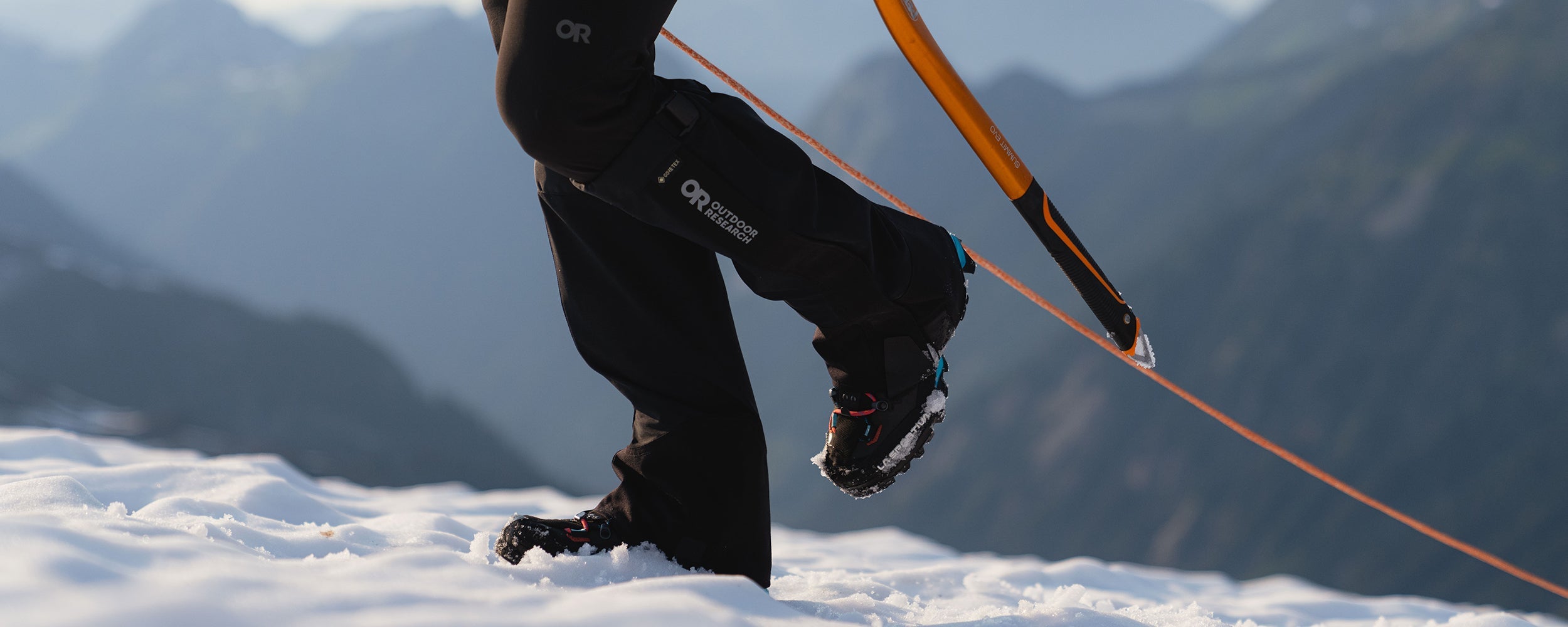 Woman wearing Outdoor Research Crocodile Gaiters in Black hikes up a snowy mountain while carrying a pick axe. 
