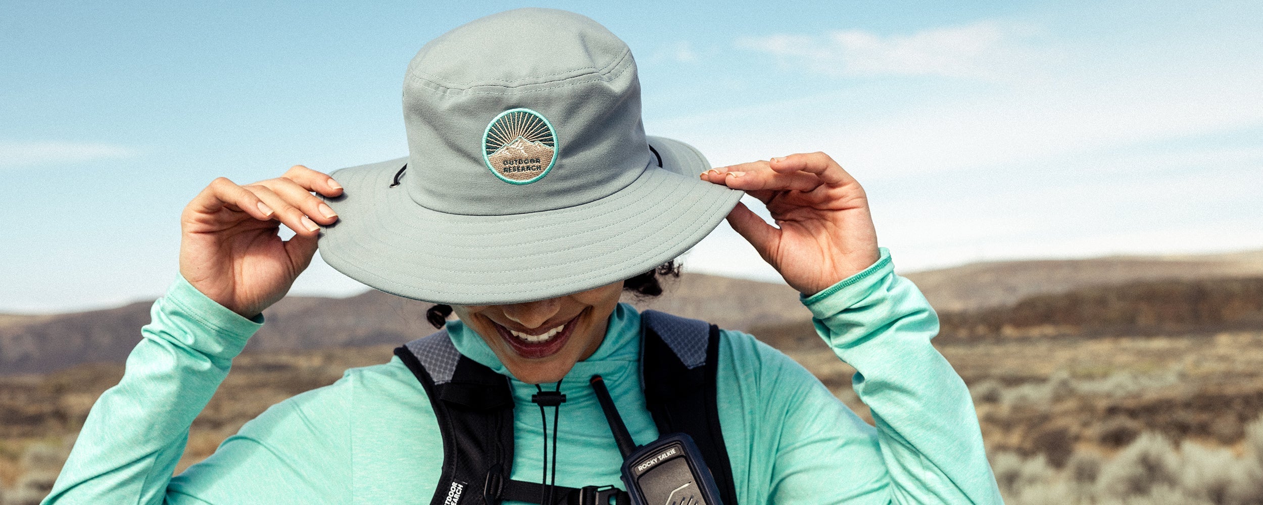 A woman smiles while grasping the sides of her Outdoor Research Heyday Boonie Hat in Neptune while out on a hike.