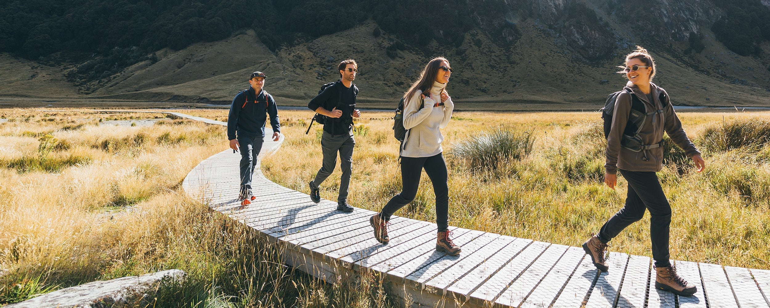 4 friends walking along a trail in the mountains wearing Outdoor Research Women's Crescent Fleece Full Zip Hoodie Sable, Women's Trail Mix XT Fleece Cowl Oyster, and Men's Trail Mix XT Fleece Half Zip Atlantic. 