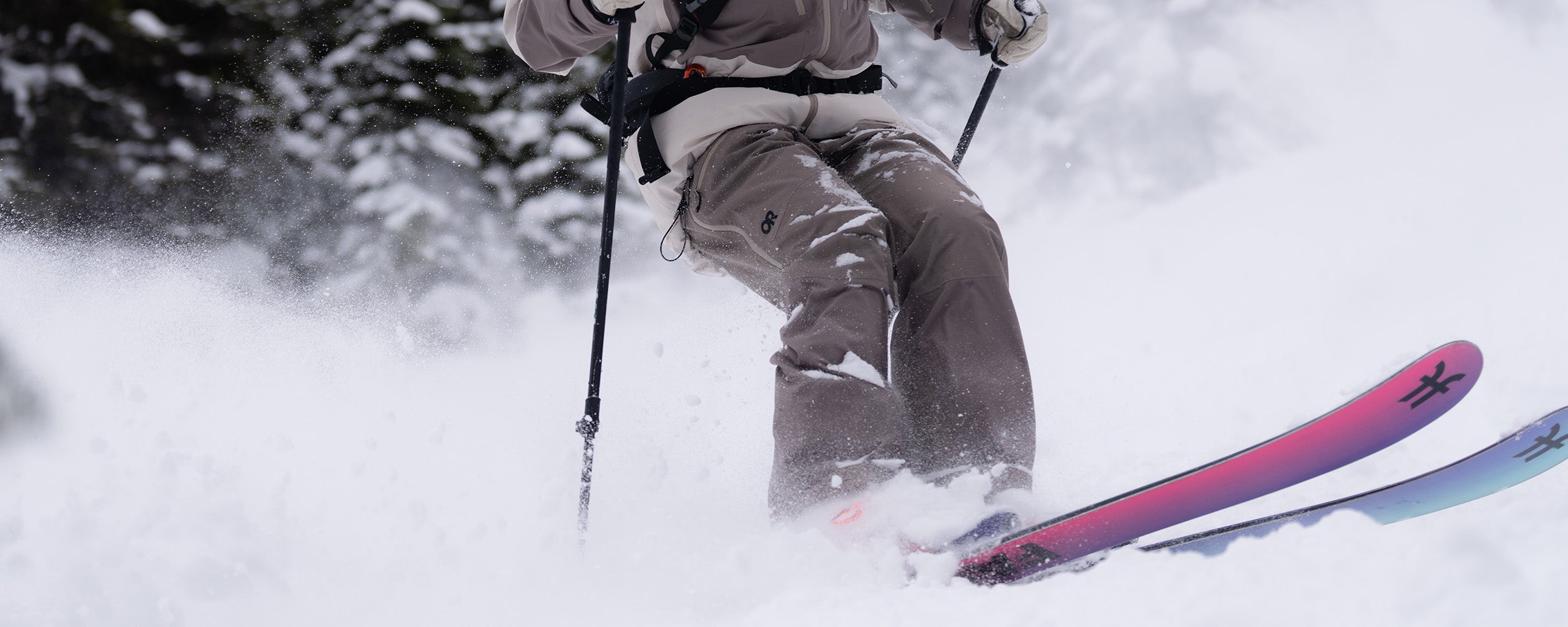 Female skiing with trees in the background wearing Outdoor Research Women's Powderverse Jacket Oyster/Sable and Women's Powderverse Pants Sable. 