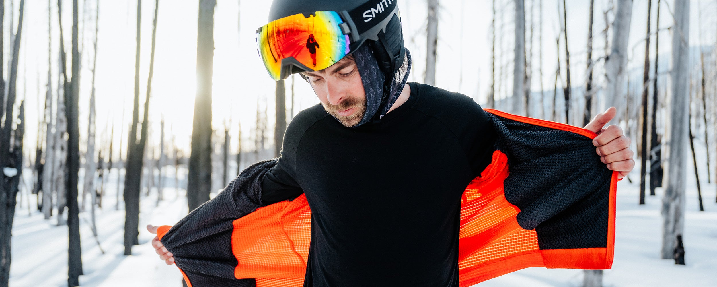A man pulls a jacket on over his black base layer top while out in the snow.
