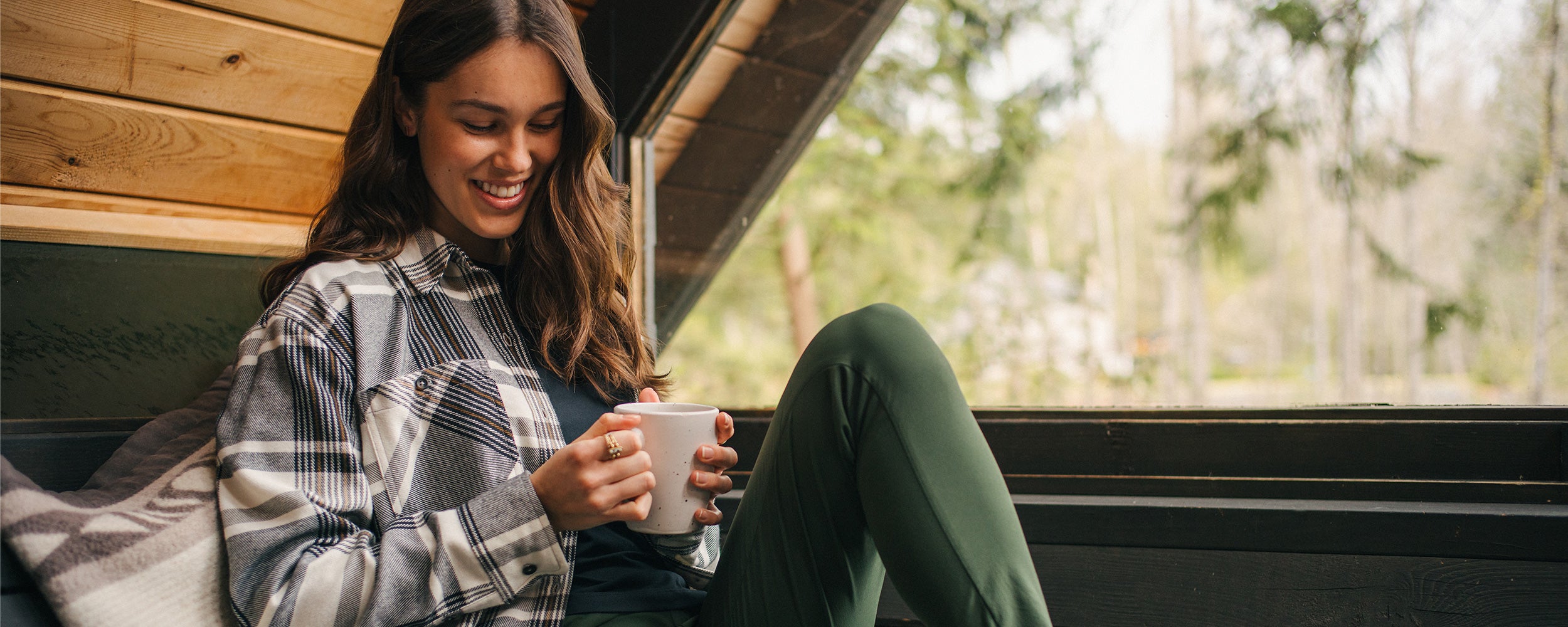A woman in a plaid flannel shirt smiles while holding her coffee in a cabin.