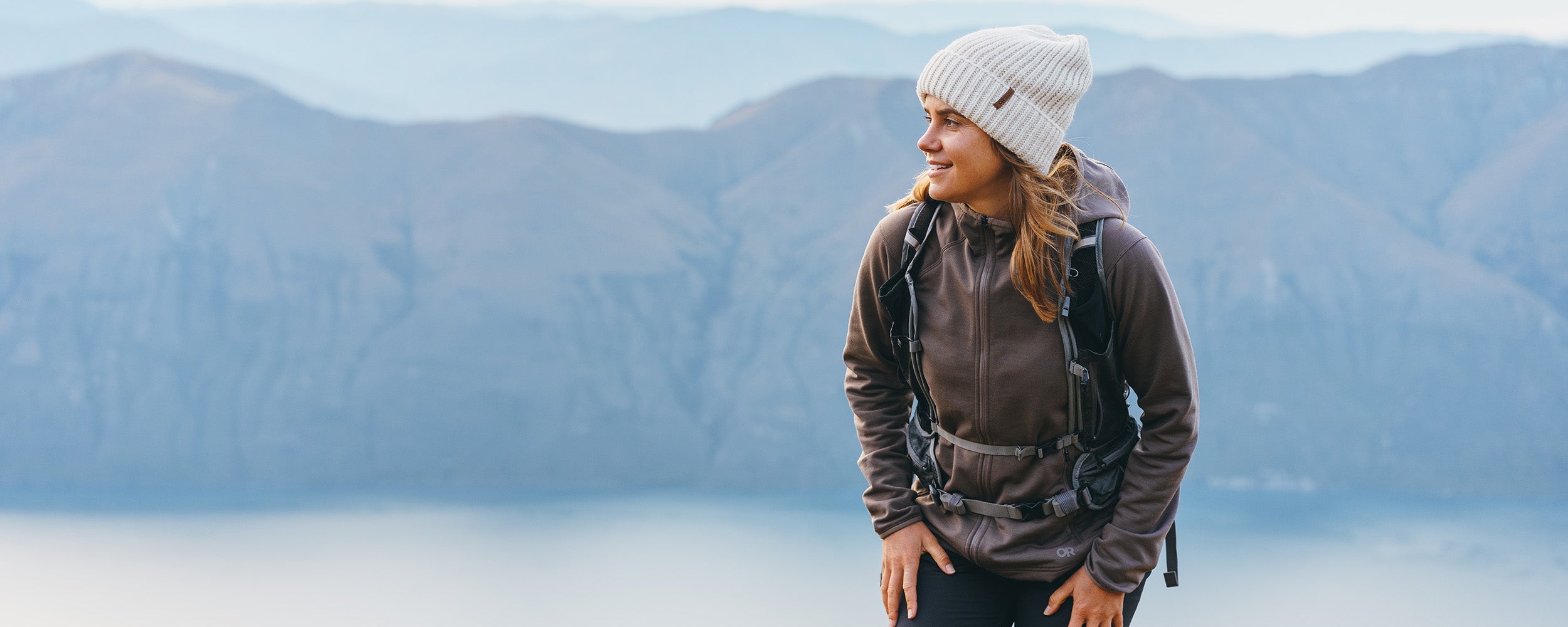 Female hiking in the mountains above the water wearing Outdoor Research Women's Crescent Fleece Full Zip Hoodie Sable and Liftie VX Beanie Sand.