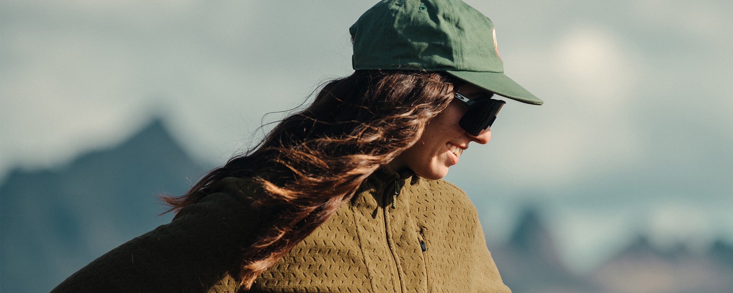 Female smiling in the sun with hands on her hips wearing Outdoor Research Women's Trail Mix XT Fleece Pullover Hoodie. 