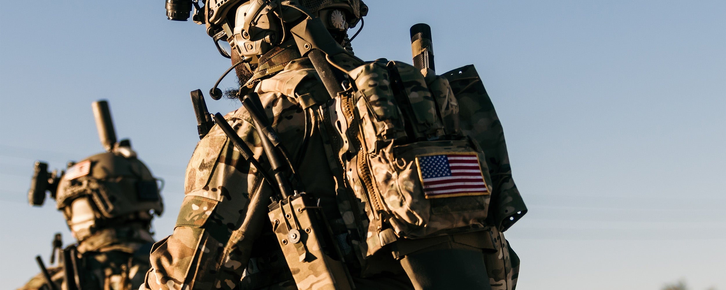 Close up of two men wearing tactical clothing and gear, with an American flag attached to one of their backpacks.