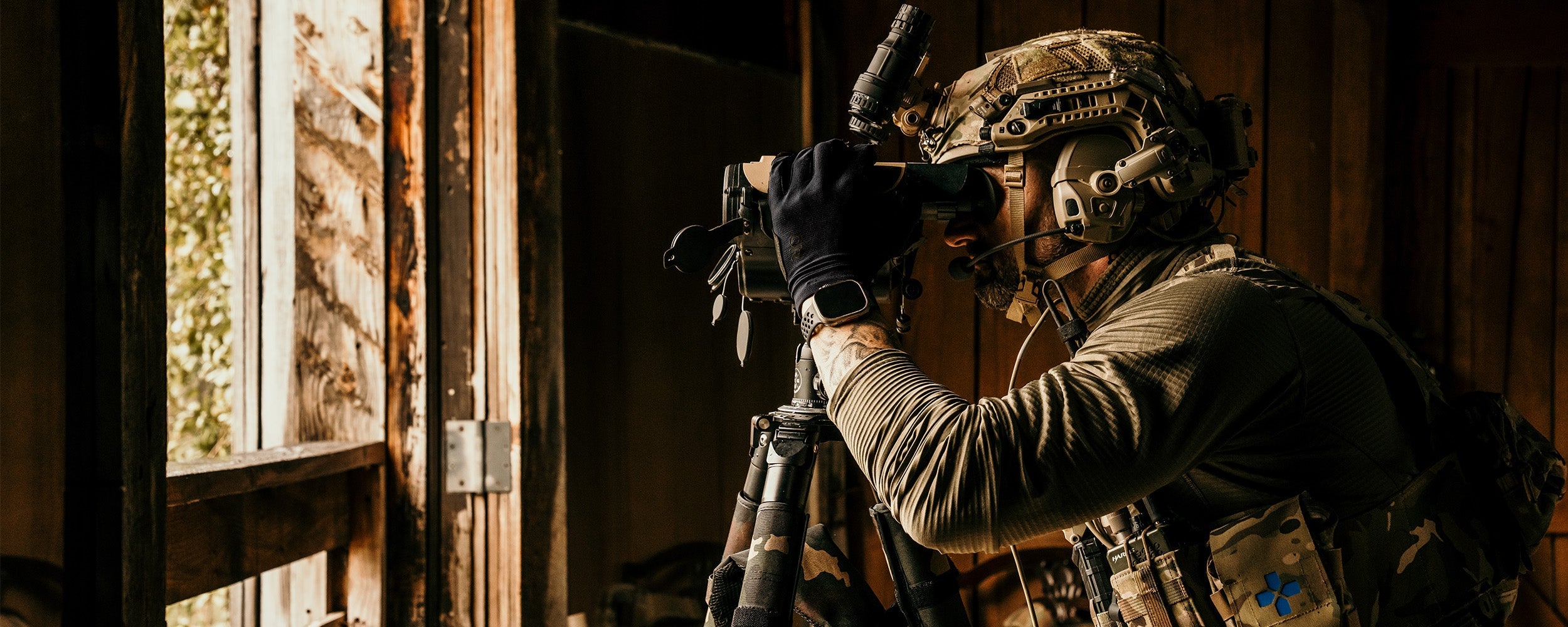 A person wearing Outdoor Research Tactical clothing looks through binoculars while standing near an open window.