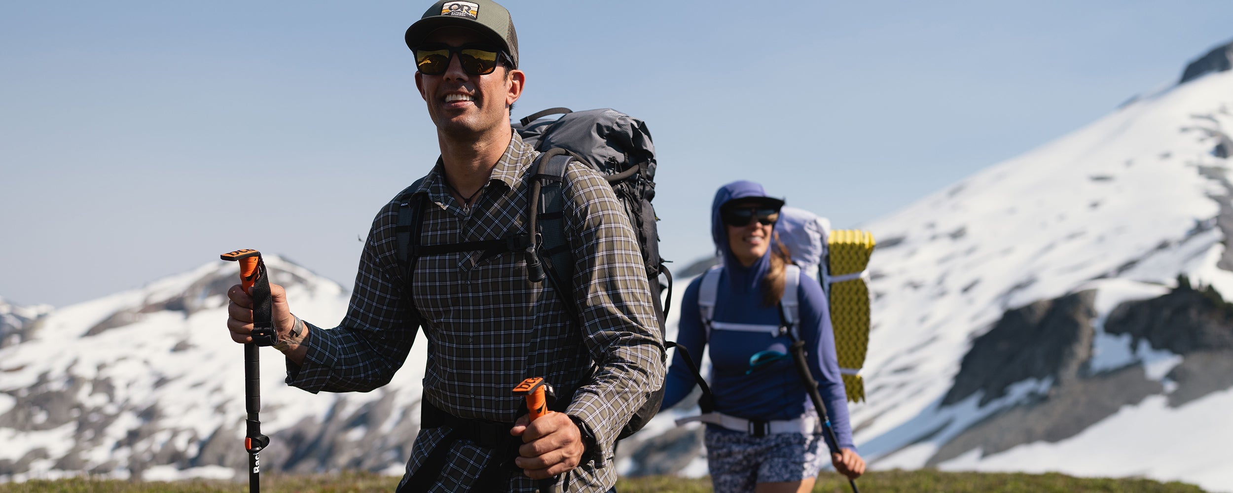 Two friends hike along a path with a blue sky and snowy mountain behind them while wearing Outdoor Research Sportswear.