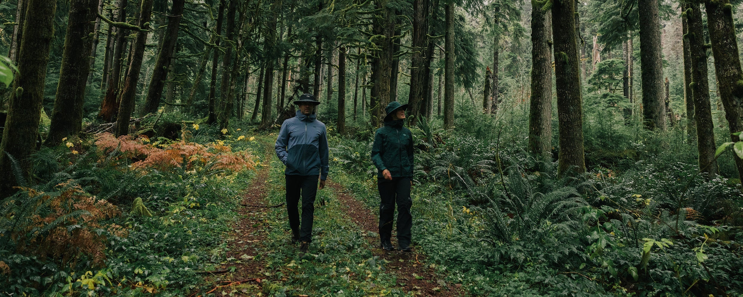 Two friends stroll through a grass path surround by tall green trees while wearing Outdoor Research Outerwear.