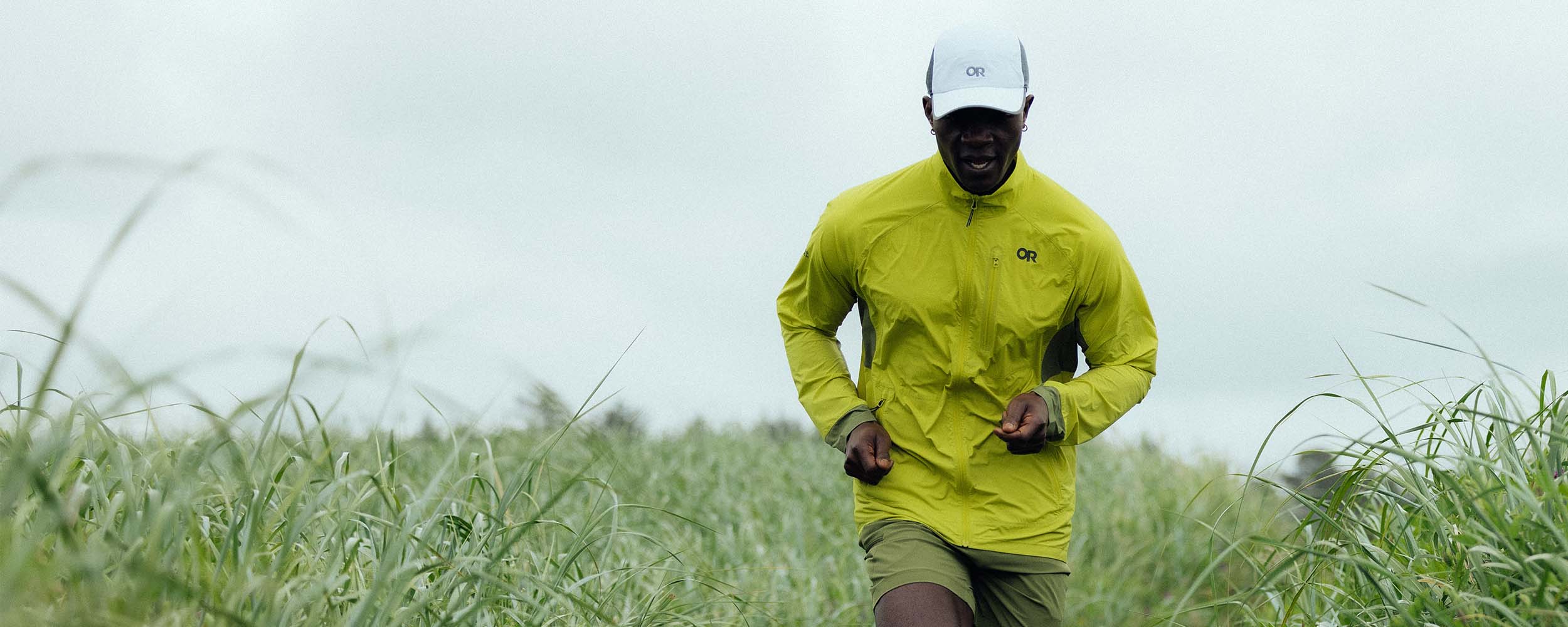 A man runs quickly along a path cutting through a tall grass field wearing Outdoor Research Deviator Wind Jacket in Viper/Willow.