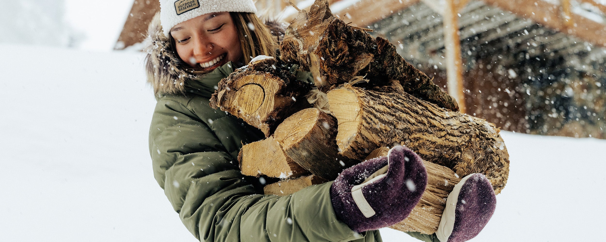 Zoe Atkin wears the Outdoor Research Coze Lux Down Parka in Ranger Green while carrying fire wood in the snow. 
