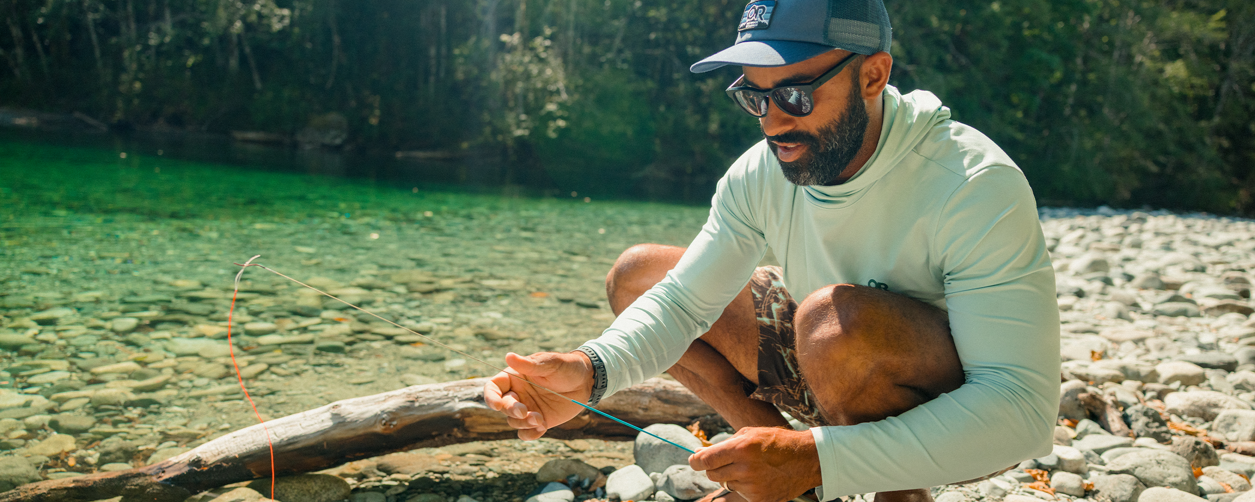 Man by the water adjusting fishing pole wearing Outdoor Research ActiveIce Spectrum Sun Hoodie Tidal. 