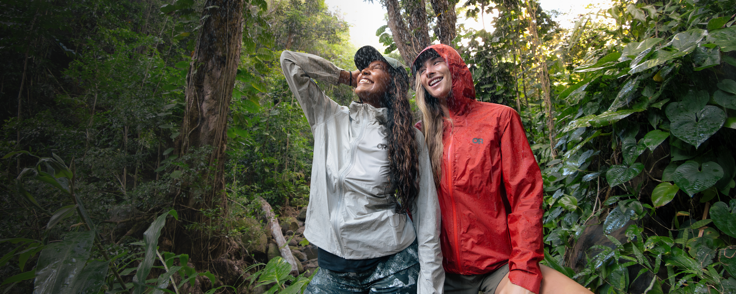 Two females in the rain wearing Outdoor Research Women's Helium UL Jackets Madrone Red and Oyster. 