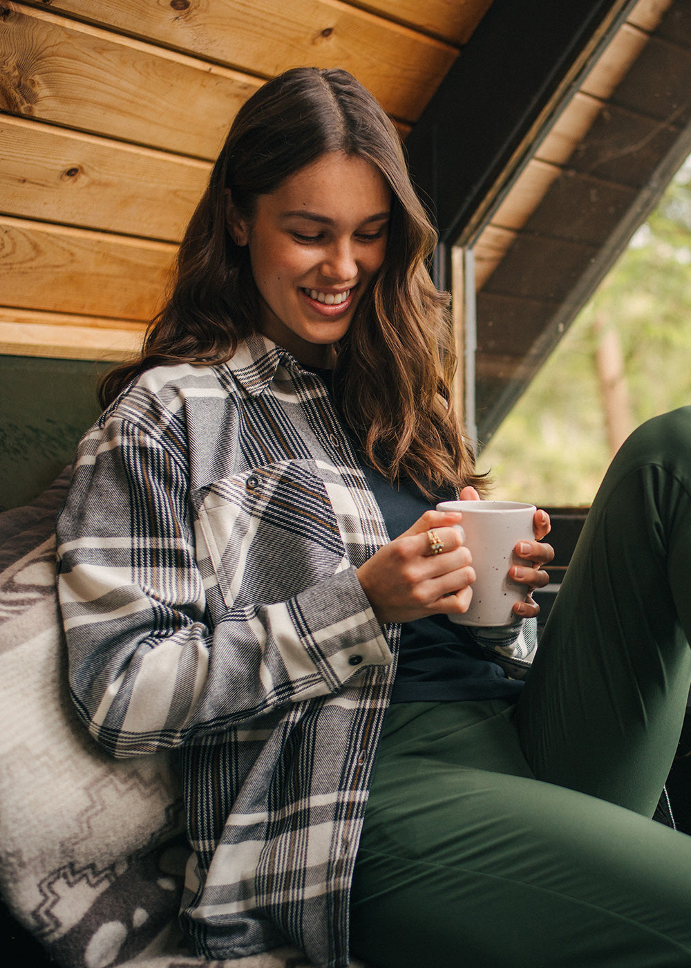 A woman in a plaid flannel shirt smiles while holding her coffee in a cabin.