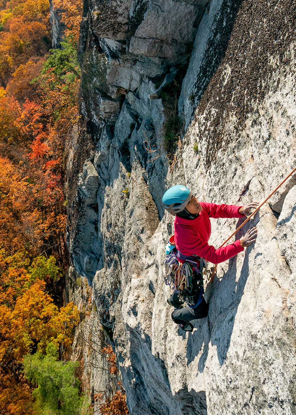 A woman climbs a rock wall with fall colored leaves on the trees below.