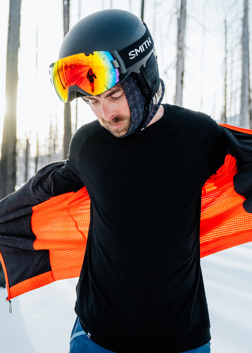A man pulls a jacket on over his black base layer top while out in the snow.