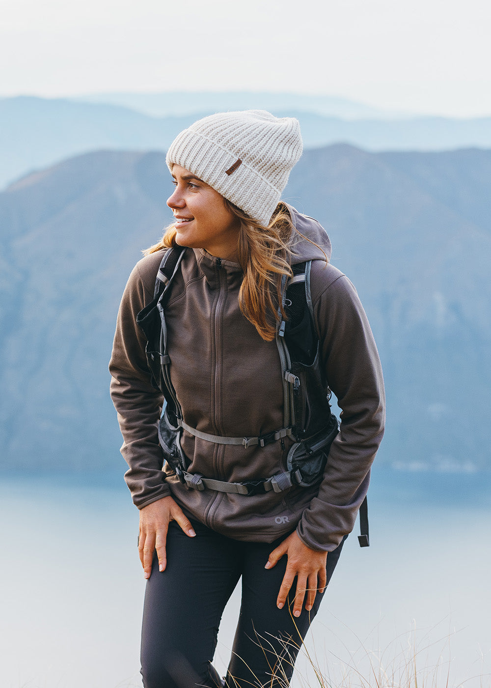 Female hiking in the mountains above the water wearing Outdoor Research Women's Crescent Fleece Full Zip Hoodie Sable and Liftie VX Beanie Sand.
