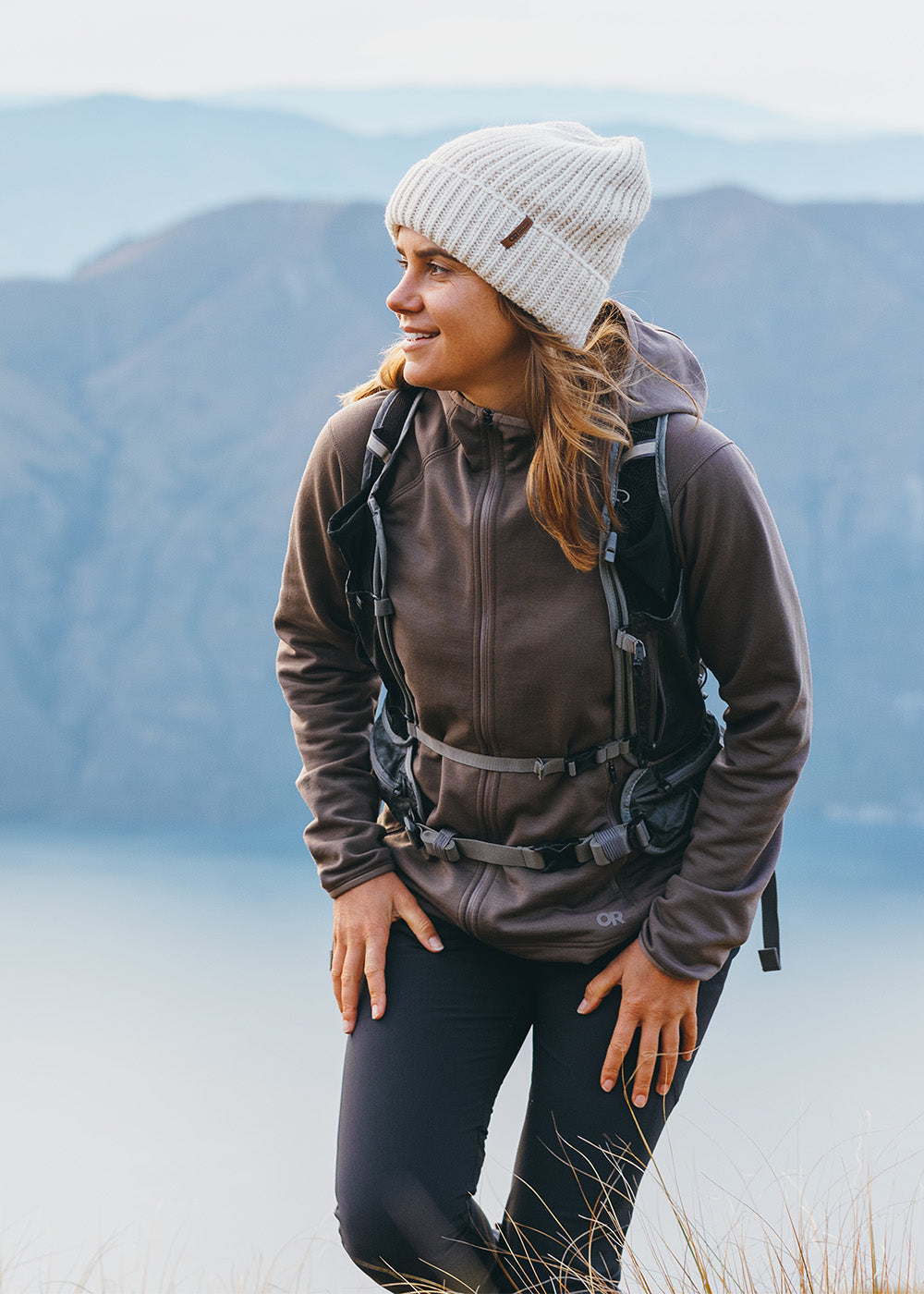 Female hiking in the mountains above the water wearing Outdoor Research Women's Crescent Fleece Full Zip Hoodie Sable and Liftie VX Beanie Sand.