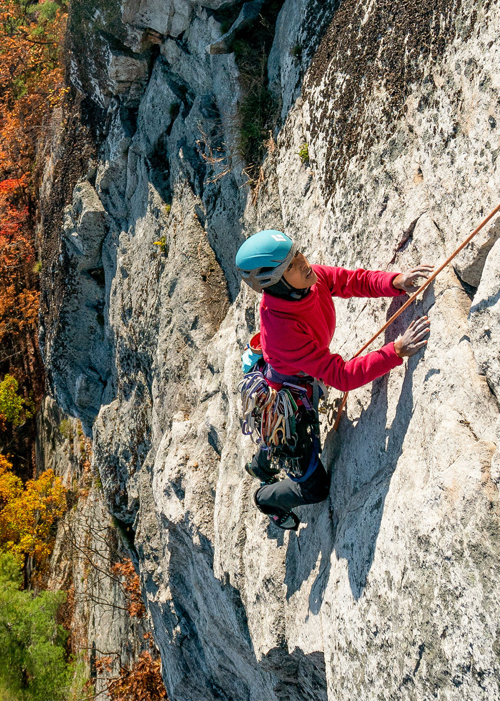 Person rock climbing on a rocky cliff face with autumn foliage in the background.