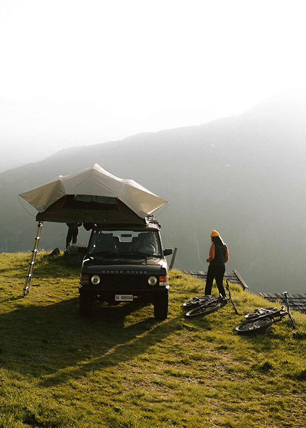 A woman walks around her truck camper on a green hillside.