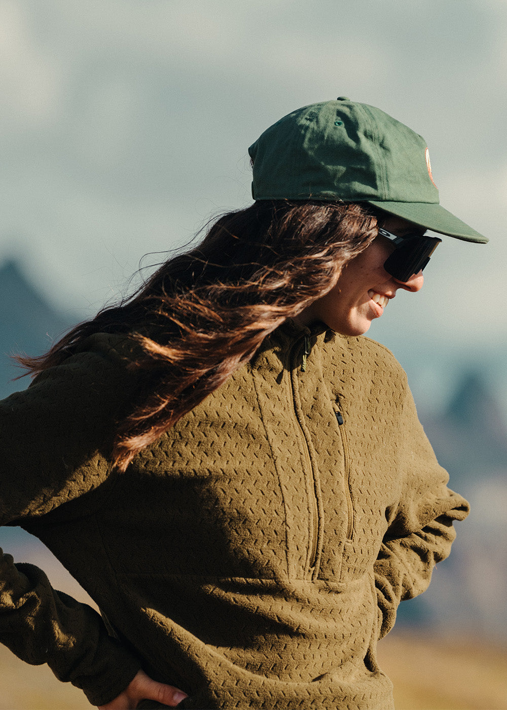 Female smiling in the sun with hands on her hips wearing Outdoor Research Women's Trail Mix XT Fleece Pullover Hoodie. 