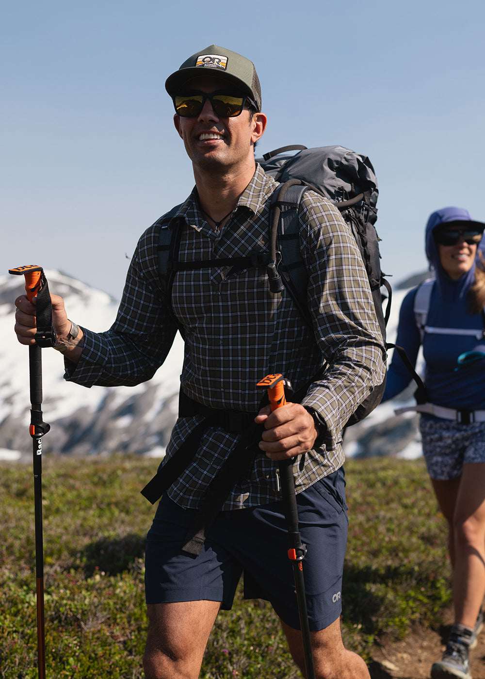 Two friends hike along a path with a blue sky and snowy mountain behind them while wearing Outdoor Research Sportswear.