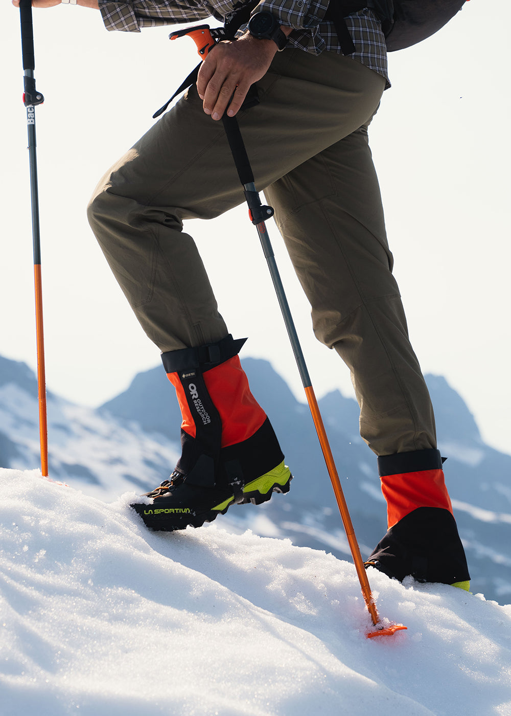  Man climbs up a snowy hill while wearing the Crocodile Mid-Height Gaiters in Spice/Black. 