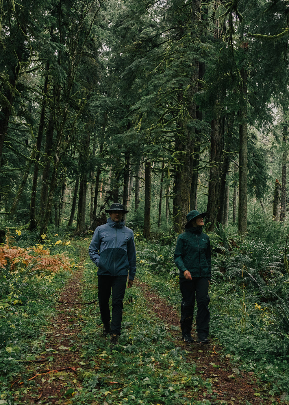 Two friends stroll through a grass path surround by tall green trees while wearing Outdoor Research Outerwear.