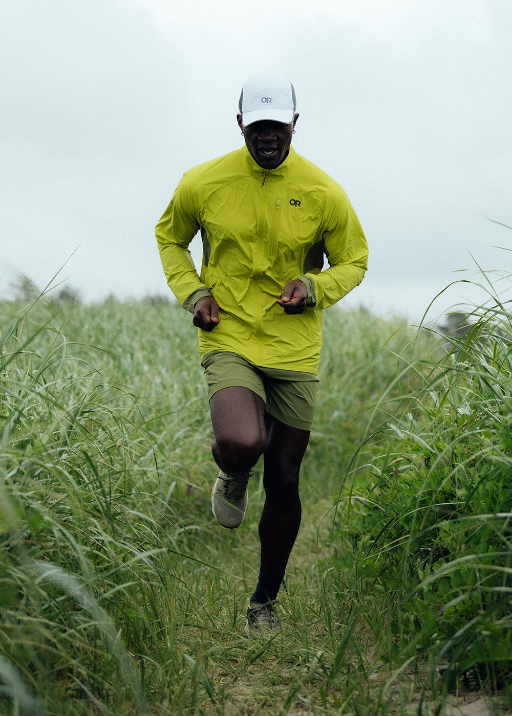 A man runs quickly along a path cutting through a tall grass field wearing Outdoor Research Deviator Wind Jacket in Viper/Willow.