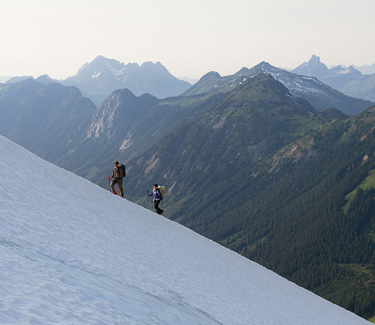 Two friends wearing Outdoor Research outerwear climb alongside a snowy mountain beneath a blue sky. 