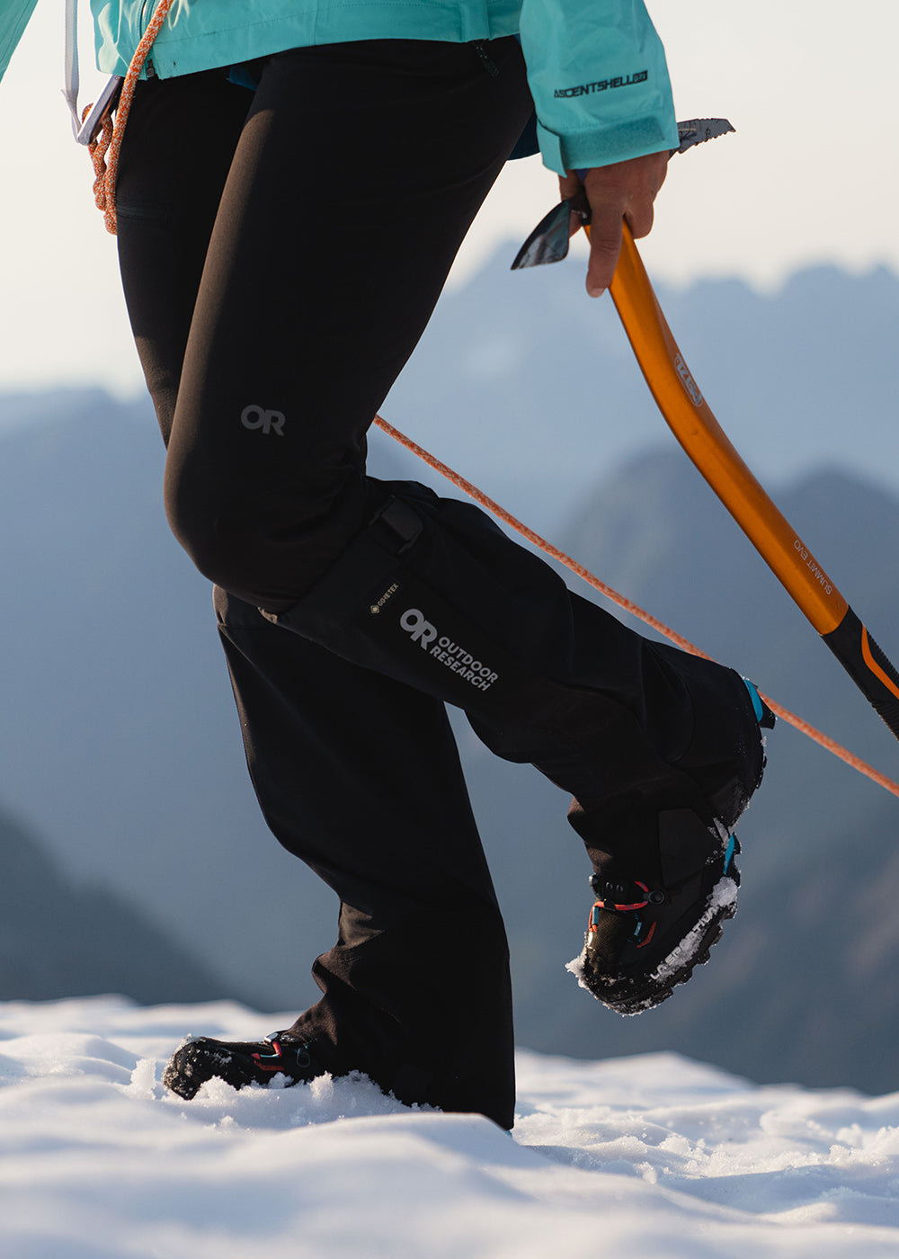 Woman wearing Outdoor Research Crocodile Gaiters in Black hikes up a snowy mountain while carrying a pick axe. 