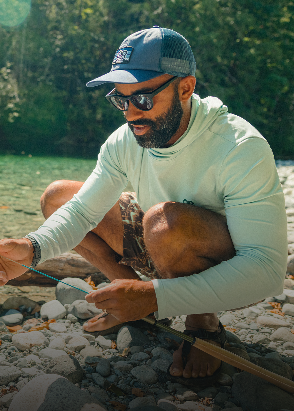 Man by the water adjusting fishing pole wearing Outdoor Research ActiveIce Spectrum Sun Hoodie Tidal. 